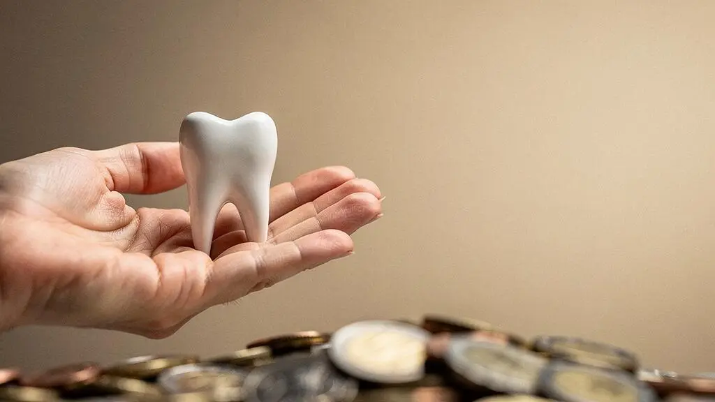 Une main tenant délicatement une dent stylisée en céramique entourée de pièces de monnaie floutées, symbolisant le financement d'un implant dentaire